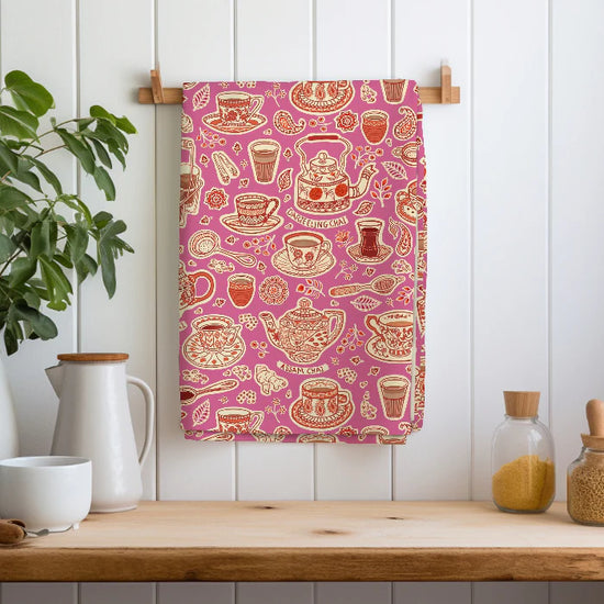 Tea-themed tea towel with teapots, cups, and spiceson a pink background hanging on a wooden rack.