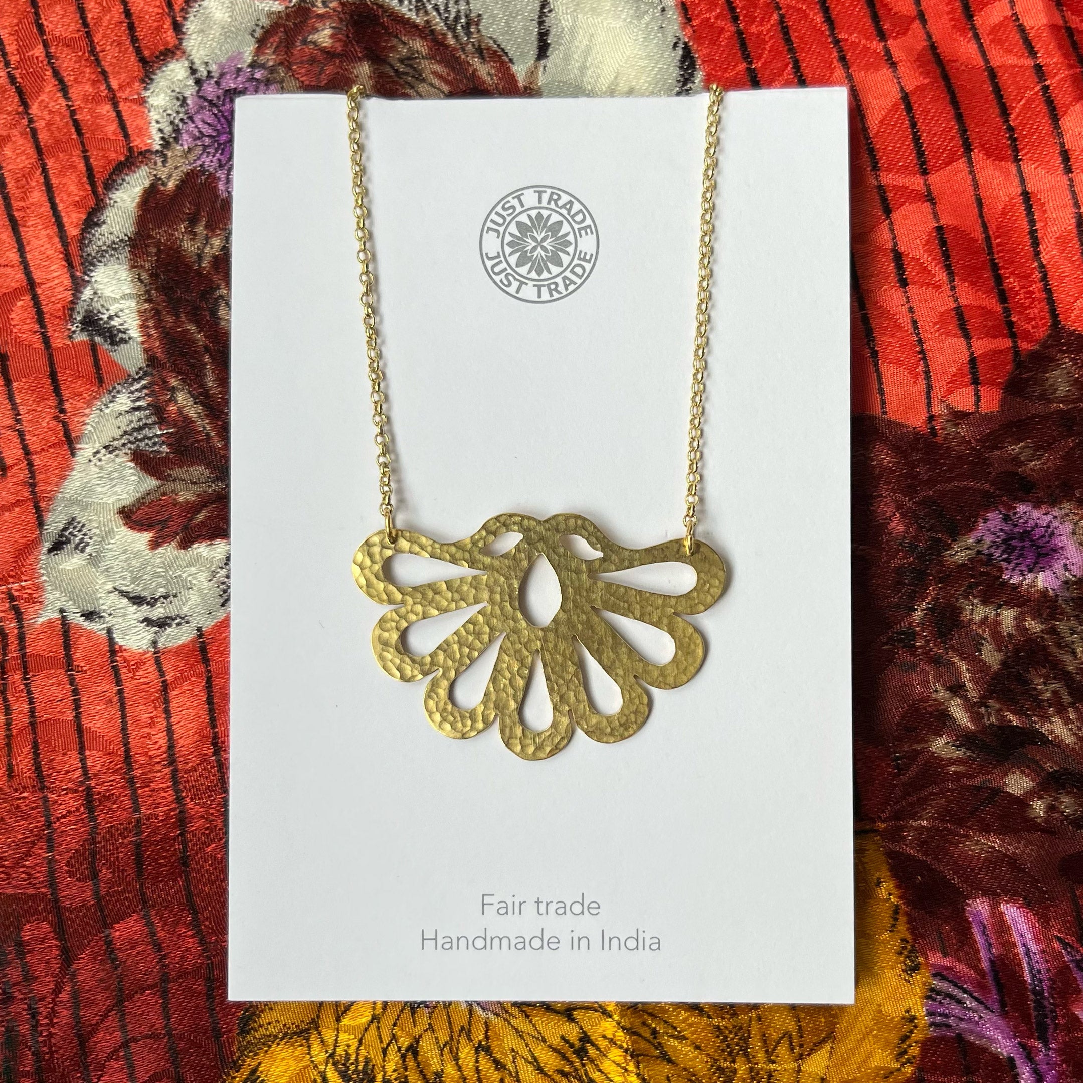 Lobed and cut out necklace pendant shaped like an upside down peacock tail. Presented against a colourful fabric backdrop.
