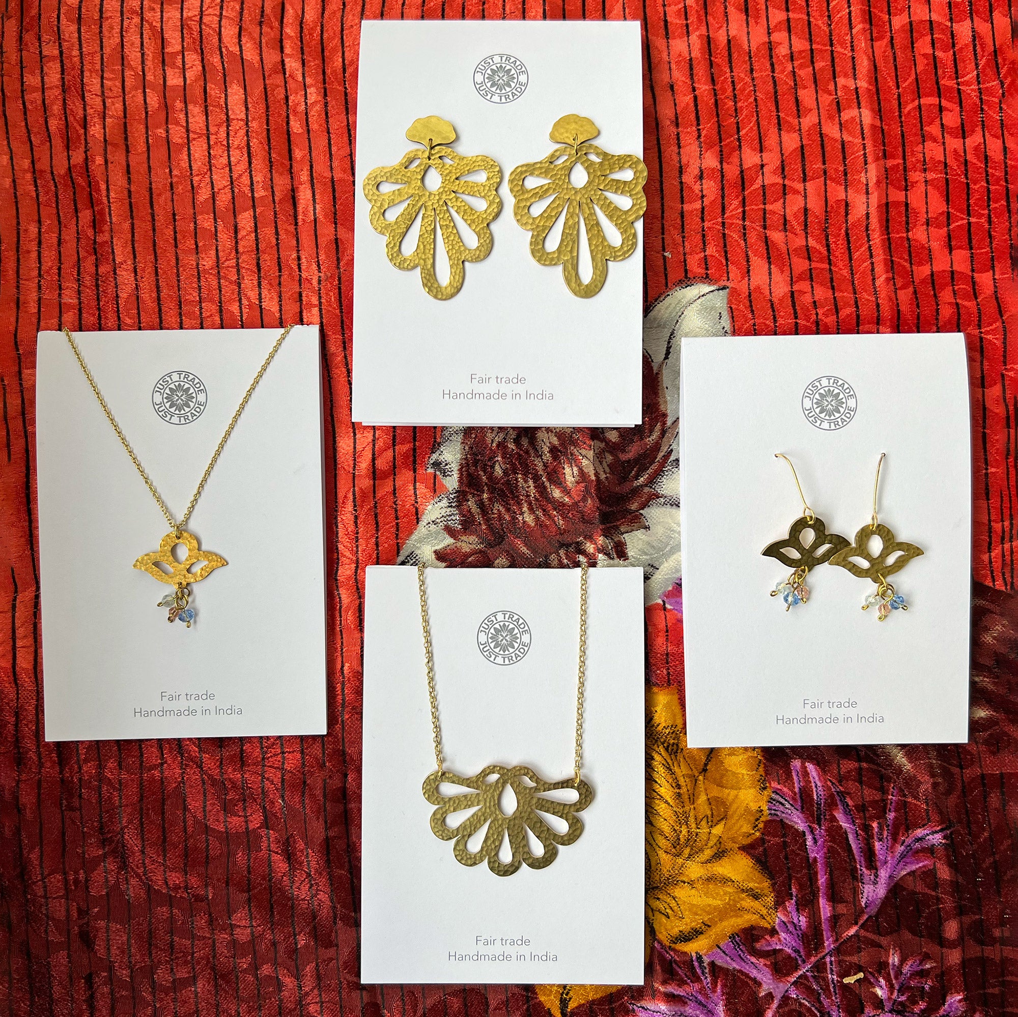 Four pieces of gold-coloured fair trade jewellery, including two necklaces and two pairs of earrings, displayed on white cards with the Just Trade logo, against a colourful patterned fabric background.