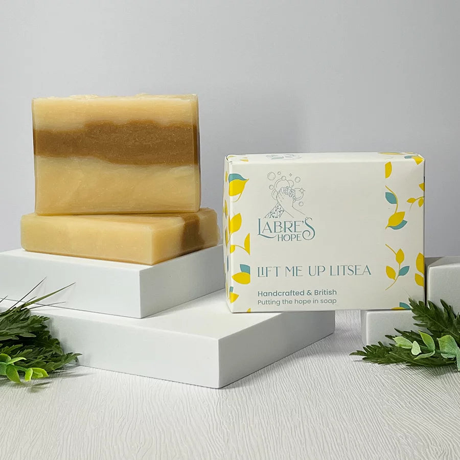 The image shows two bars of soap stacked on top of each other. The soap has a layered appearance with a light yellow color at the top and a darker brown color at the bottom. The soap is packaged in a white box with a label. There are also some green leaves and white boxes in the background. 