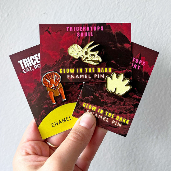 Hand holding glow-in-the-dark enamel pins with packaging against a white background