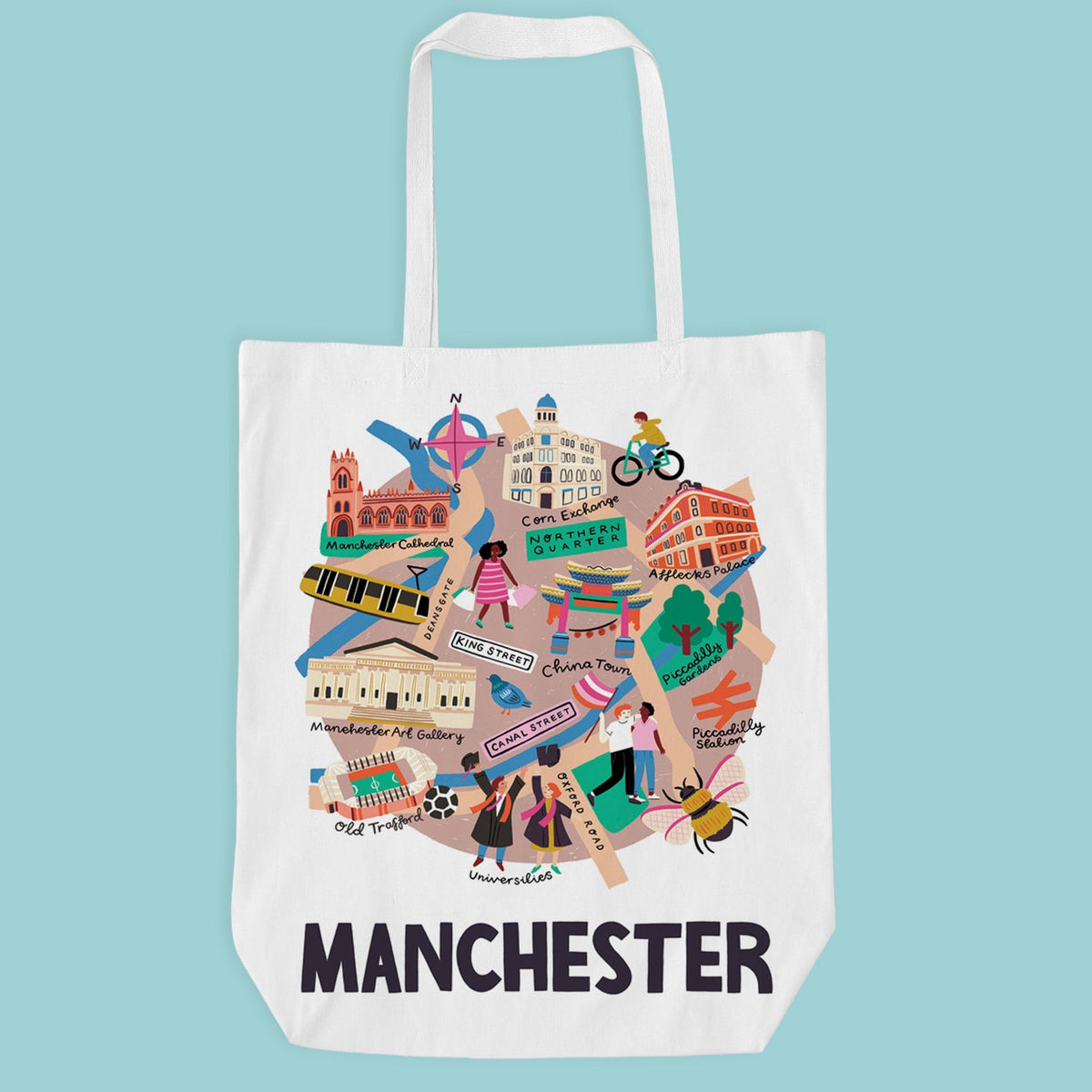 A white canvas tote bag featuring a colorful illustrated graphic of Manchester landmarks, street signs, and people, with the word 'MANCHESTER' printed in bold black letters below.