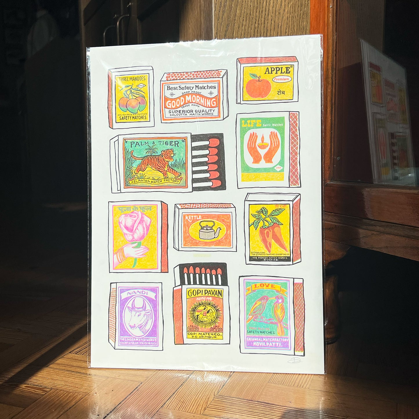 A colourful print showing lots of different matchboxes with cool pictures and names on them, like "Three Mangoes" and "Palm & Tiger".