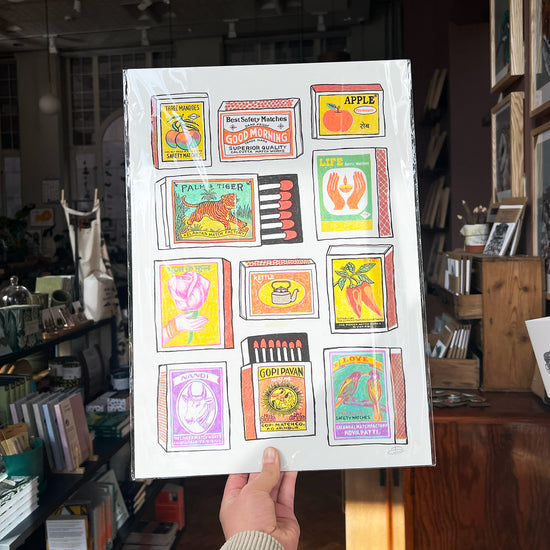 Someone holding up a colourful print showing lots of different matchboxes with cool pictures and names on them, like "Three Mangoes" and "Palm & Tiger".