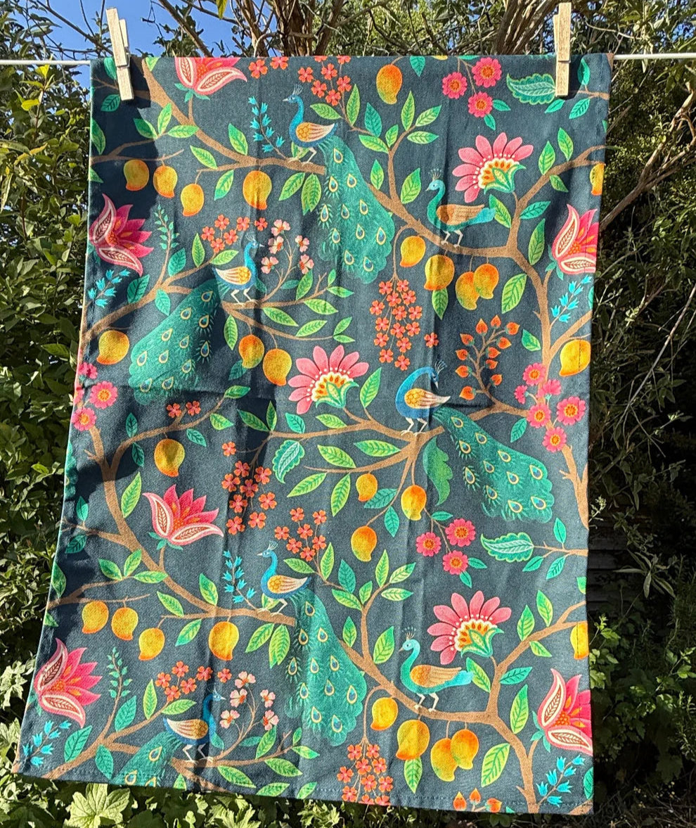 Colorful tea towel with a peacock in a mango tree repeating motif hanging outdoors on a line with greenery in the background.