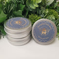 Two round silver metal tins of Springfield Park Bees honey, one stacked and one standing, with blue lids featuring a bee logo and honeycomb pattern, set against green artificial pine branches and leaves.