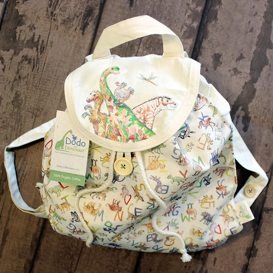 Lifestyle photo of the cotton rucksack seen from slightly above. The rucksack has a buttom to hold the top flap down and cords from the internal closure can be seen trailing out from under the flap. Dinosaurs and a dodo motifs on the flap and in smaller on the body of the bag is an extinct animal alphabet in a mish-mash repeat pattern.