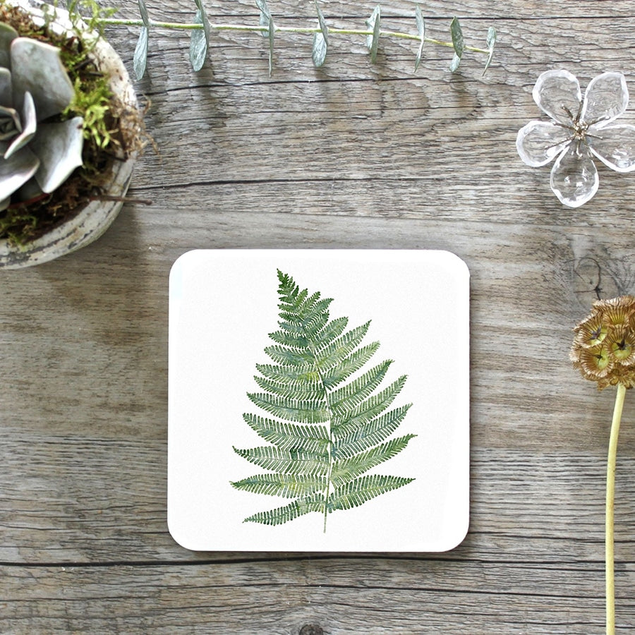Square coaster with rounded corners and a watercolour picture of a fern plant. Photographed on a grey wooden surface.
