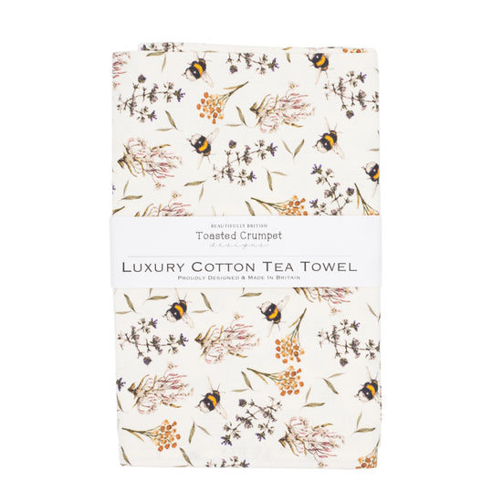 Tea towel with bee and floral pattern folded into a rectangular shape with a white belly band with Toasted Crumpet branding. Photographed against a white background.