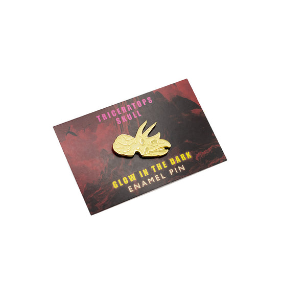 Gold enamel pin of a triceratops skull on a dark card with text