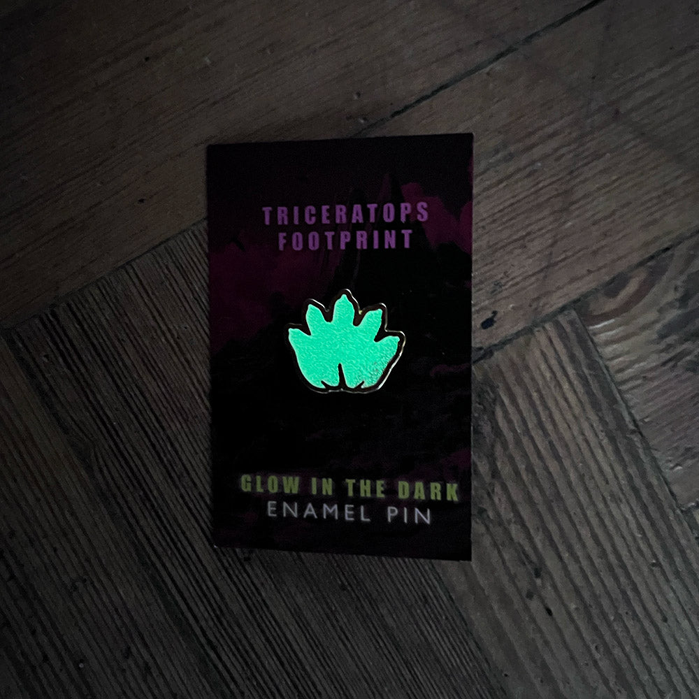 Glow-in-the-dark enamel pin with triceratops footprint design on a dark background