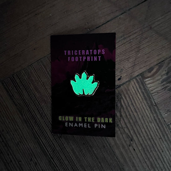 Glow-in-the-dark enamel pin with triceratops footprint design on a dark background