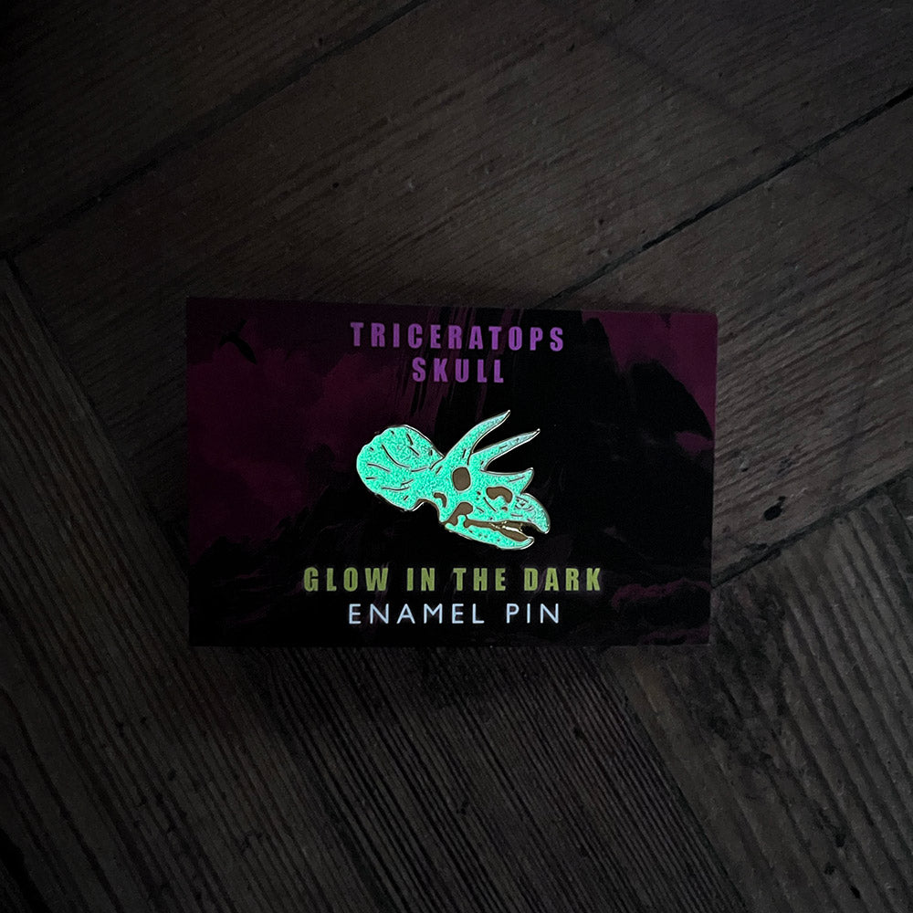 Glow in the dark enamel pin packaging with triceratops skull design on a dark wooden surface.