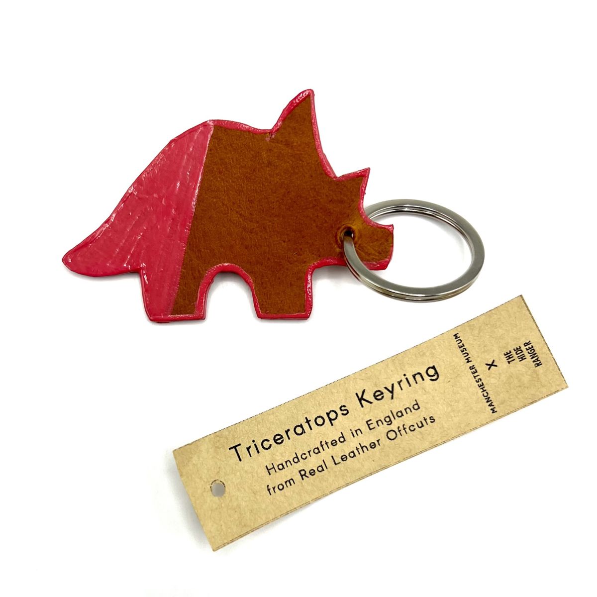 A Triceratops shaped keyring. The tail and half of the hind leg is painted pink and the same pink is painted as an outline around it's body. The keyring is where the eye would be and a paper swing tag is fastened to the keyring with a black safety pin. Tag text: Triceratops keyring. Handcrafted in England from real leather offcuts.