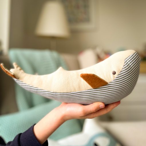 lifestyle of whale toy with stripy fabric belly and beige topside. The whale is held on the palm of a small hand.