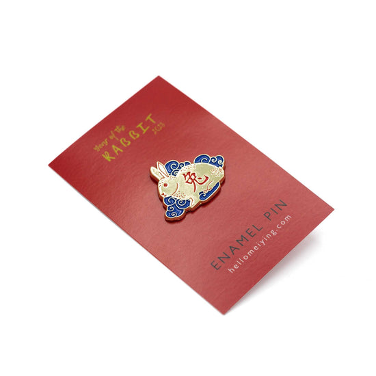 Red card backing at a slight angle with the golden and blue rabbit for year of the rabbit pin. The rabbit itself is golden with red chinese character in the body, it's sitting amongst blue clouds.
