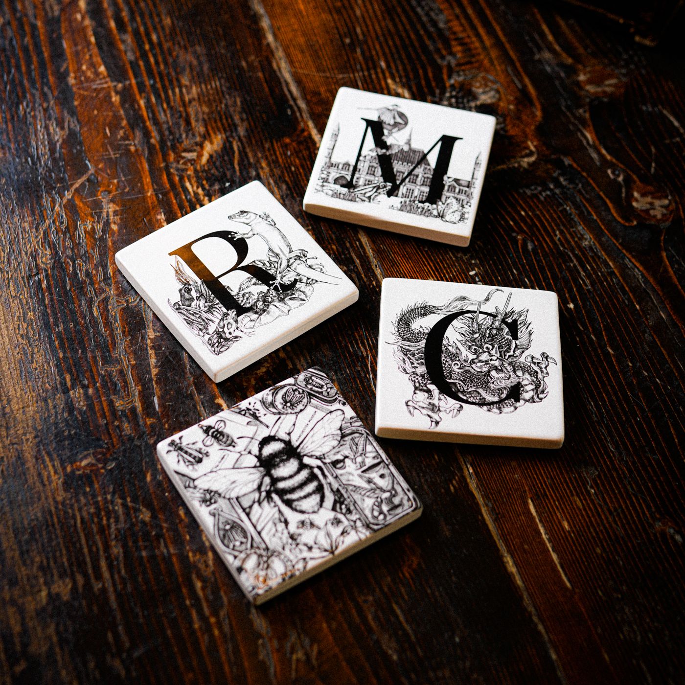 Lifestyle shot of the full four coasters from the sculpts. The M, C and R are at the top with the bee slightly out of focus near the bottom. All four coasters are resting on a dark brown wooden surface.