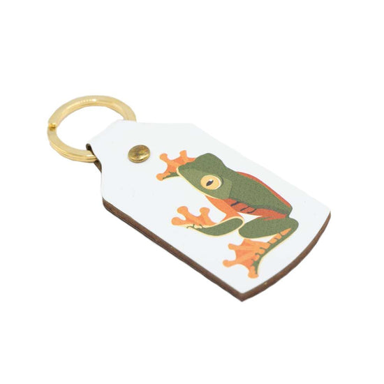 The white leather keyring lying at an angle with the green and orange frog facing opposite the point of view.