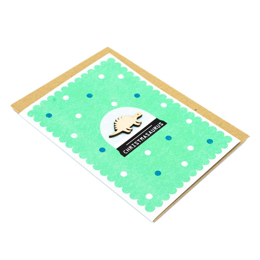 Image of a card with a brown envelope tucked inside. White background. Card is green with a wooden dinosaur decoration at the centre of a snowglobe. 