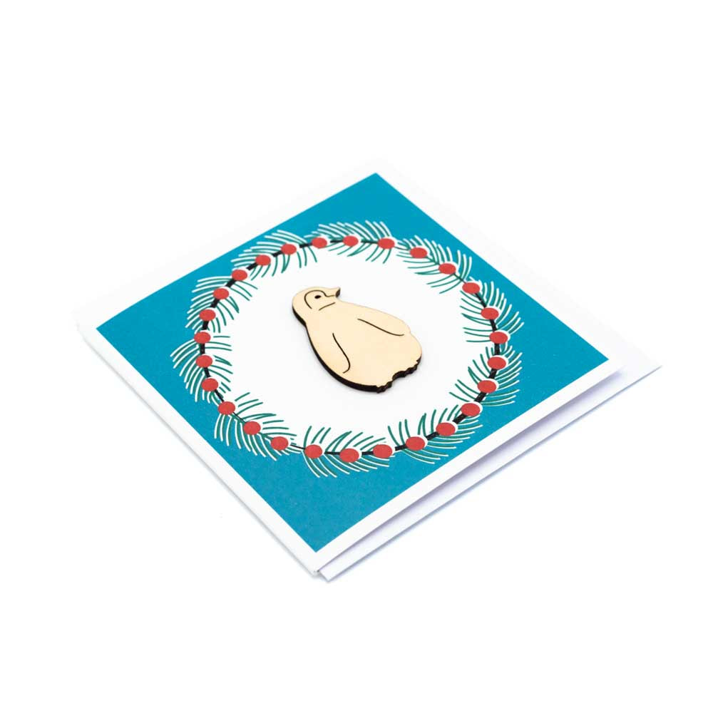 Square card with white envelope tucked inside, white background. Wooden penguin decoration at the centre of a Christmas wreath.