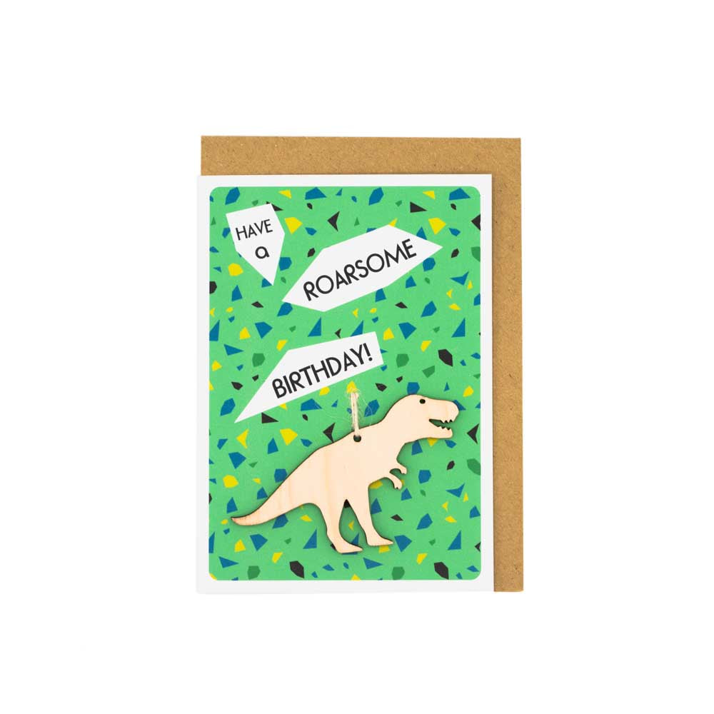 Card with white envelope tucked inside, white background. Card is green with different patterned shapes with a wooden T.Rex decoration. The words 'Have a Roarsome Birthday' above.