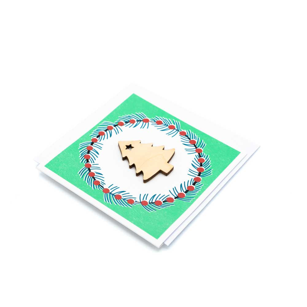 Square card with white envelope tucked inside, white background. Wooden Christmas Tree decoration at the centre of a Christmas wreath.