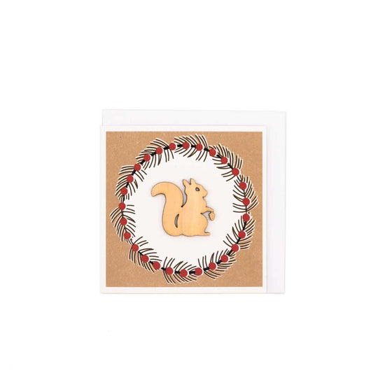 Square card with white envelope tucked inside, white background. Wooden squirrel decoration at the centre of a Christmas wreath.