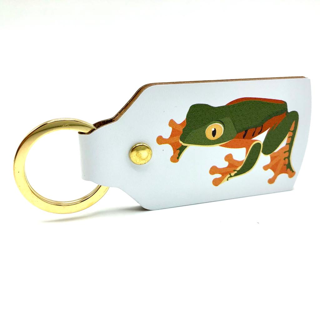 The white leather keyring standing on the narrow edge with the green and orange frog facing the left. The ring is golden.