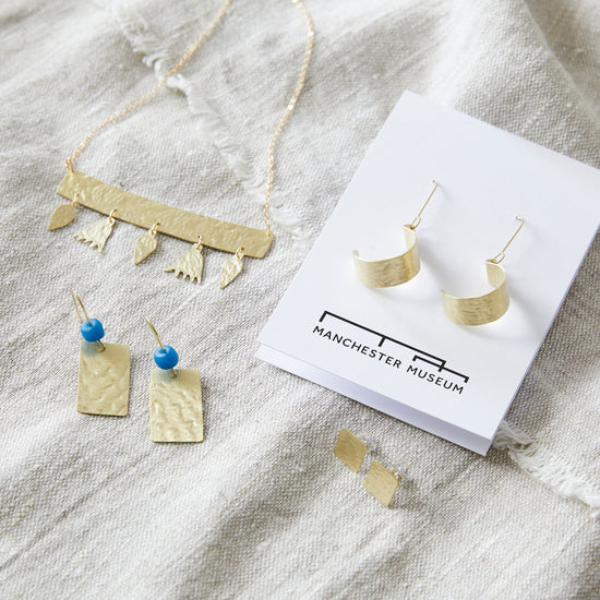 Lifestyle shot of hoop earrings on white backing card, resting on top of natural linen fabric. To the left the flower charm necklace is and below that the rectangular pendant earrings. Below/in front of the hoop earrings' backing card are the square stud earrings.