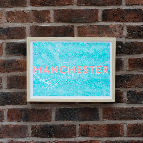 A picture of a risograph print in an ash frame. The words MANCHESTER are printed in orange above a blue and white map illustration of Manchester. Photographed against a brick wall.