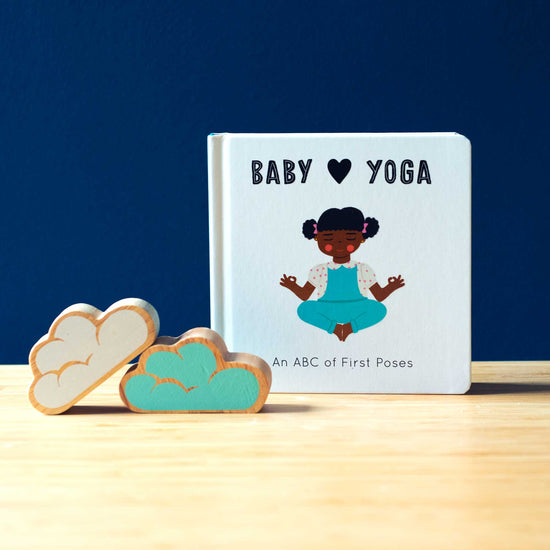 Lifestyle photograph of a book titled BABY YOGA and some cloud wooden toys, photographed on a brown table against a navy background