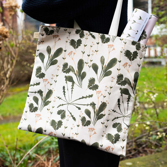 A person holding a cotton botanical print tote bag  in front of a garden