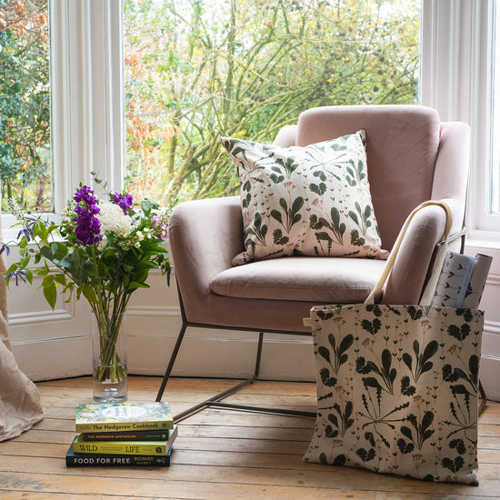 Lifestyle image of a pink chair in front of a window. On the floor by the chair is a botanical printed tote bag, a pile of books and a pot of flowers.