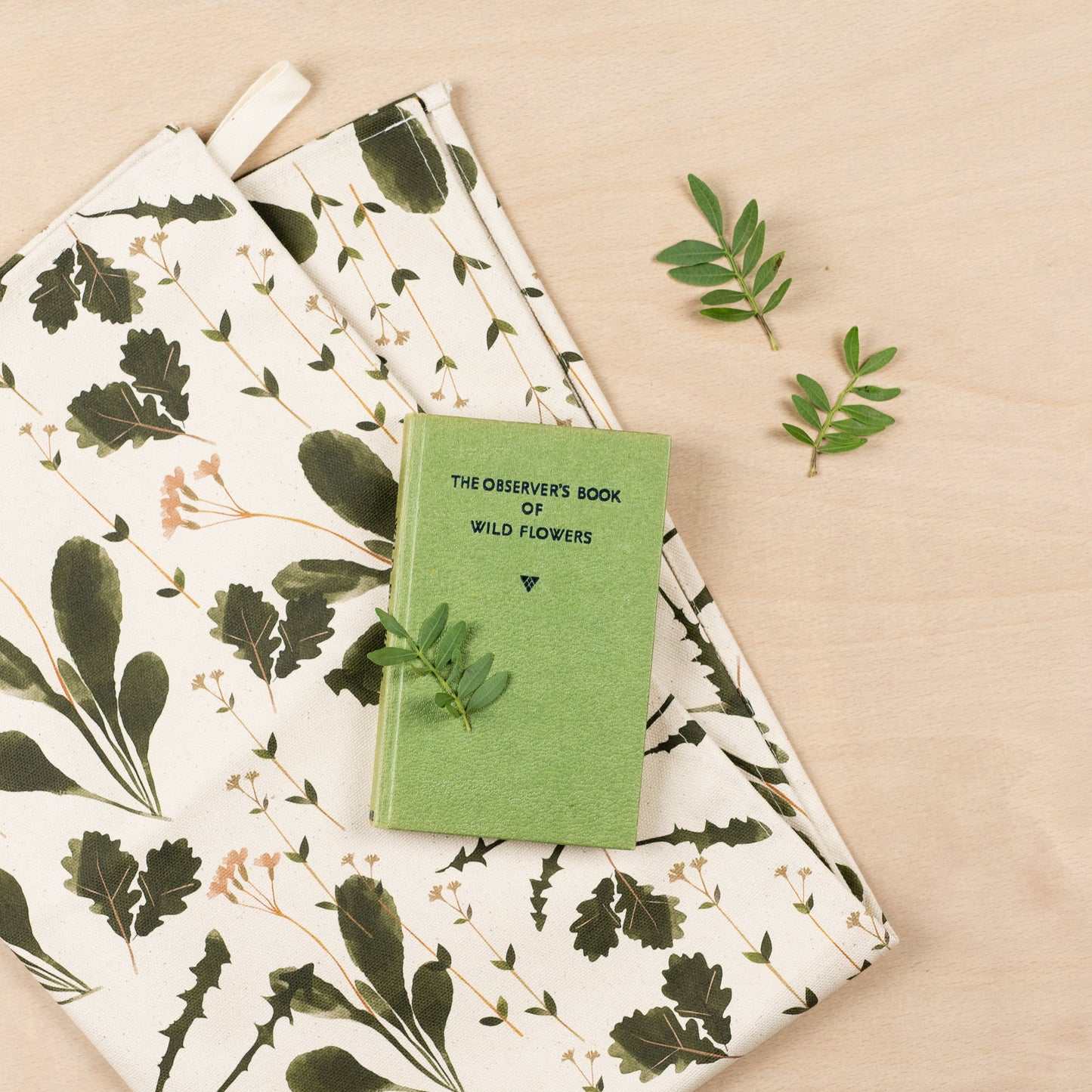 A folded tea towel on a wooden surface with a green book of wildflowers on top of the tea towel.