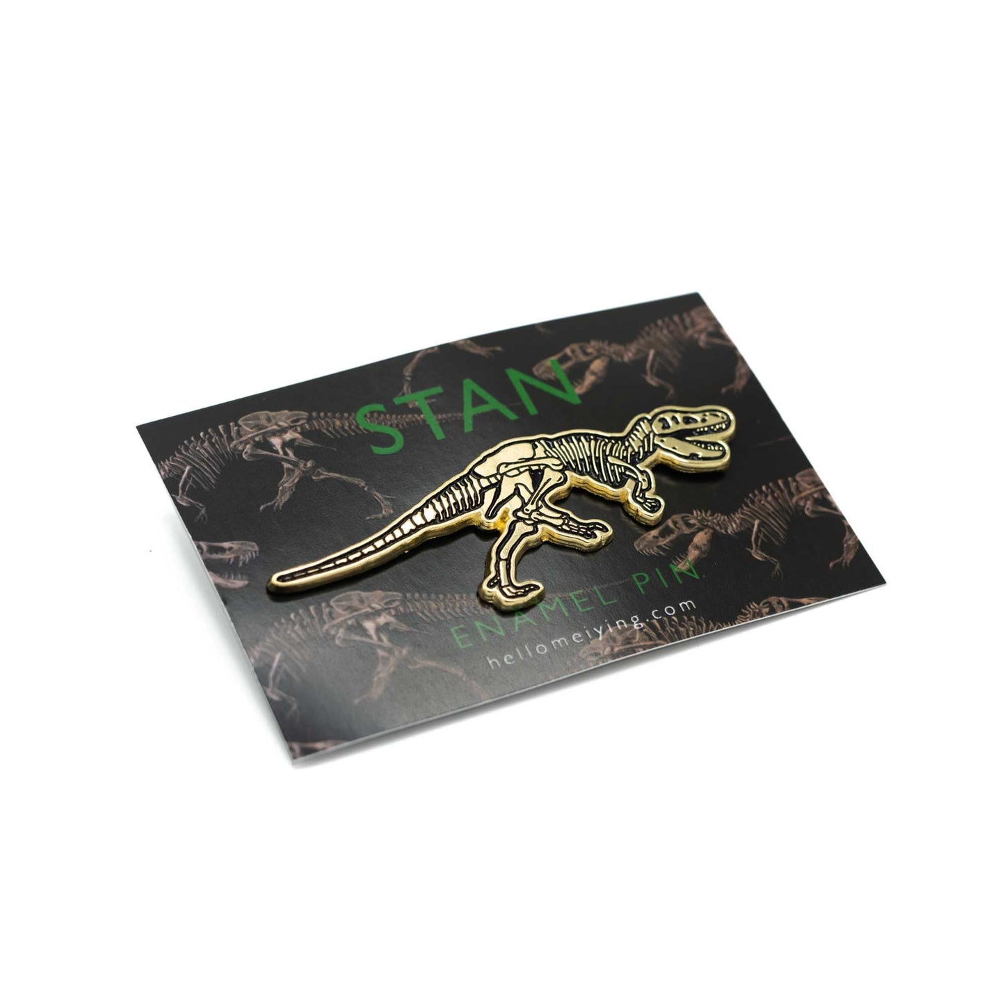 The golden Stan pin on the dark backing card seen at a slight angle which has the Stan illustration in a repeat pattern. Above the pin in dark green sans serif font it says, Stan.