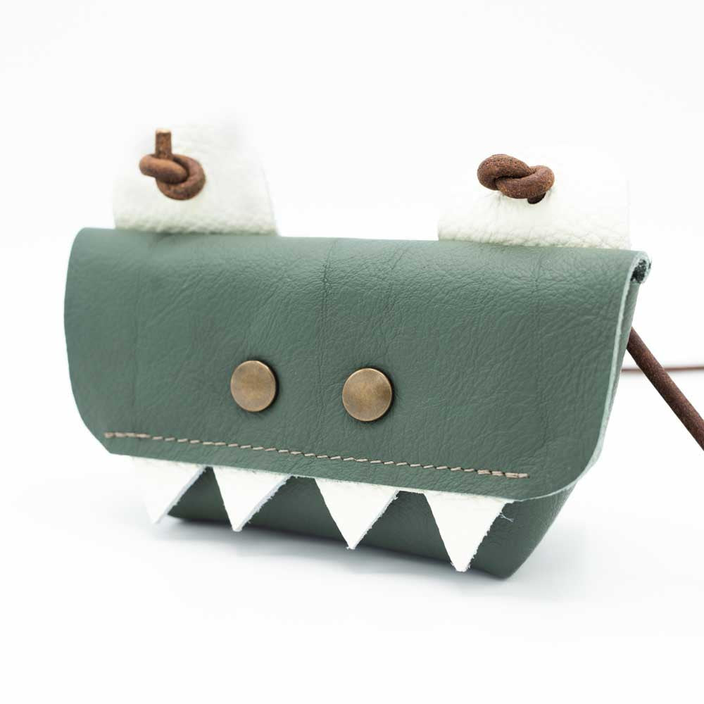 Slightly angled view of dark sage green t-rex bag with four white fangs along the  front flap of the bag.