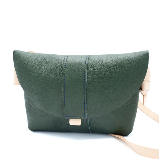 Dark green leather bag with natural pale leather strap. Seen from the front standing on a white surface.