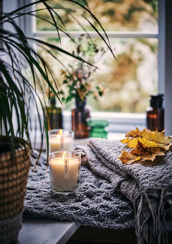 Load image into Gallery viewer, Lifestyle shot with an atmospheric lighting and autumnal leaves on a grey blanket with the lit candle in the central focus. Plants can be seen to the left and behind.
