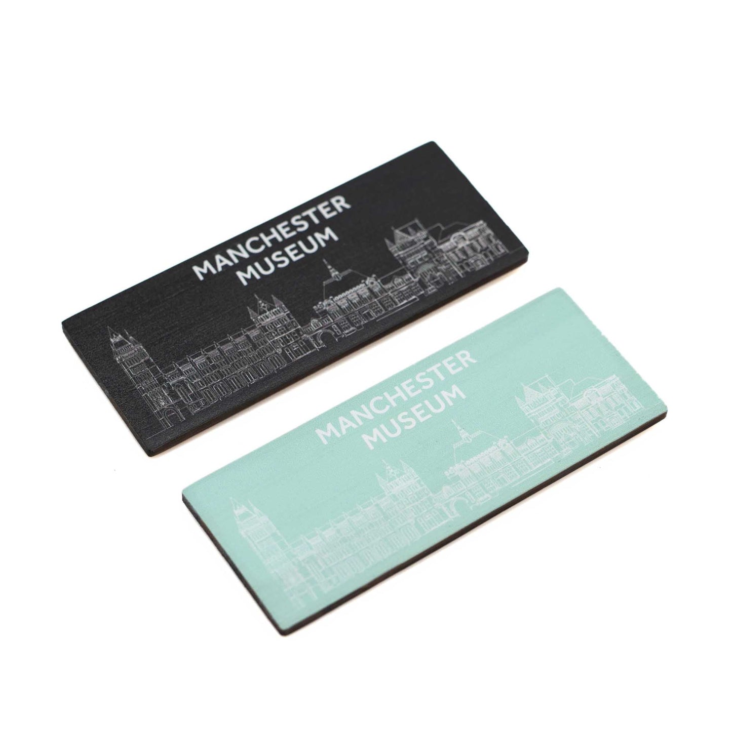 Angled view of the black and turquoise bamboo magnets on a white background. The black with white print and text is on the top. The museum facade is across the bottom of each magnet with the text, Manchester Museum, above.