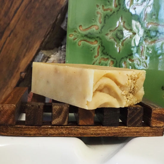 One of the beige soaps with rolled oats sprinkled on top. The soap is lying on a wooden soap holder and a green ceramic tile in the background.