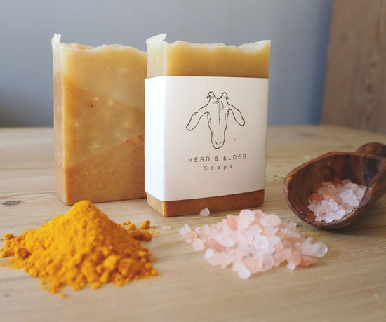 Two of the turmeric soaps standing on a wooden surface. In the foreground is a small heap of ground turmeric and Himalaya salt as well as a scoop with pink Himalaya salt.