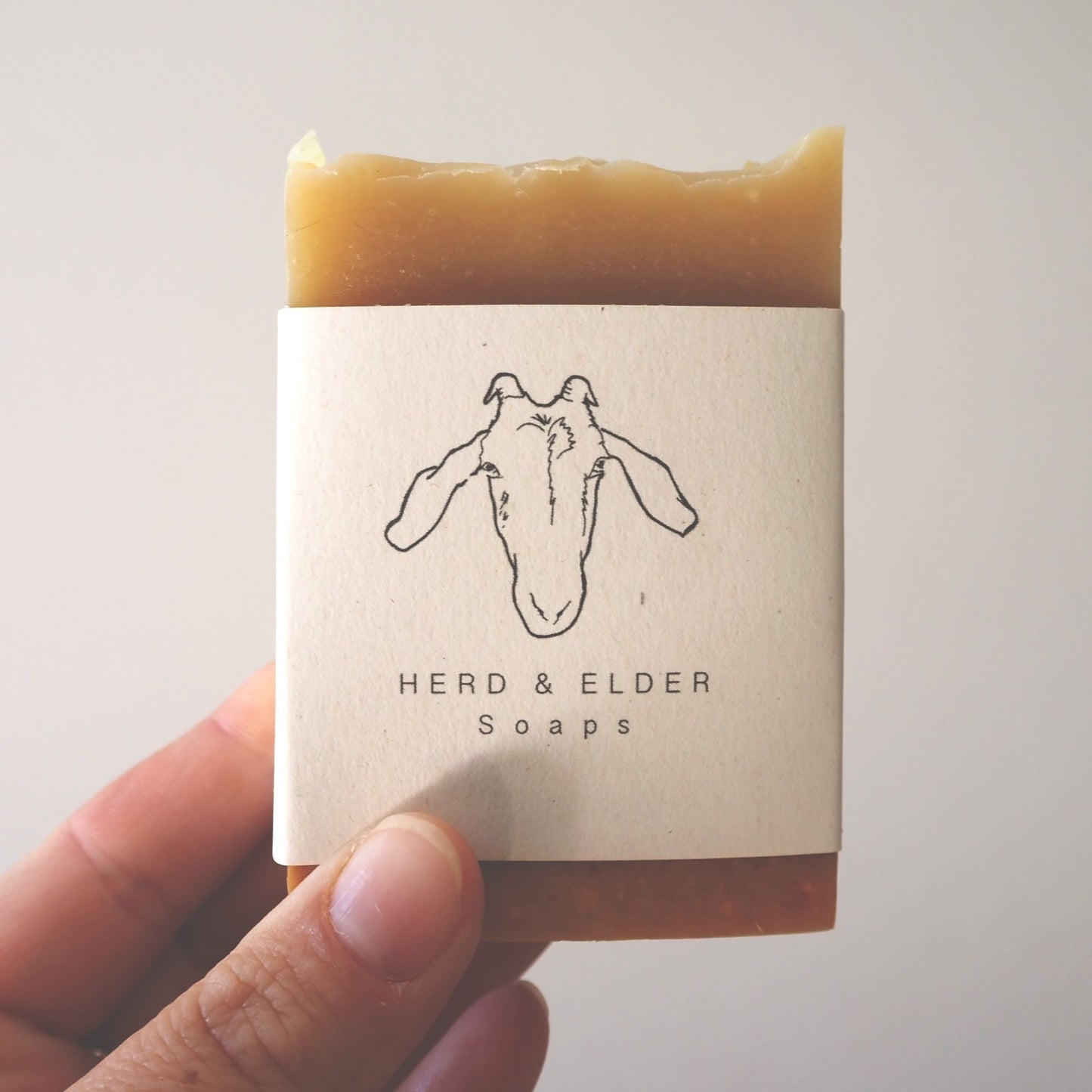 A golden soap with the herd and elder soaps branding belly band around the middle. The soap is being held up in front of a white background,