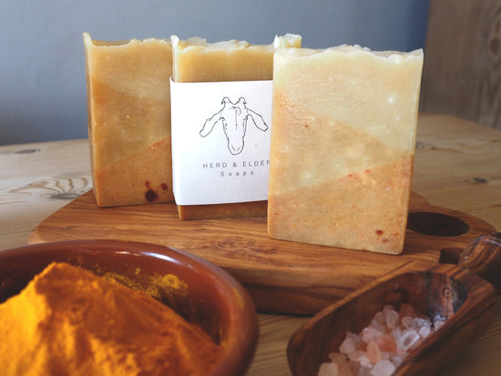 three of the turmeric soaps standing on a wooden chopping board. In the foreground is a small bowl of ground turmeric and a scoop with pink Himalaya salt.