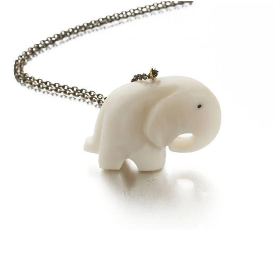 Undyed tagua pendant carved inthe shape of a small, simple elephant with the trunk rounded out and then attached to a front leg. The necklace chain is draped across the white, blank surface behind the elephant and moving out of the photos edge on the left.