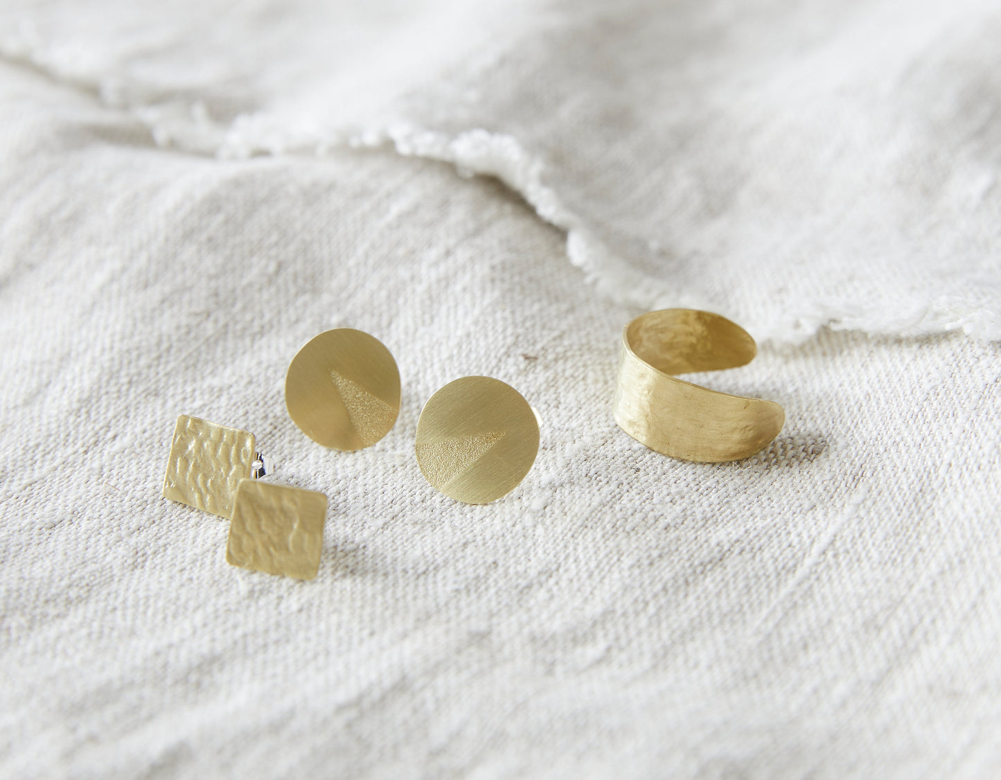 Lifestyle shot of square stud earrings, round stud earrings and the textured ring all resting on natural linen fabric.