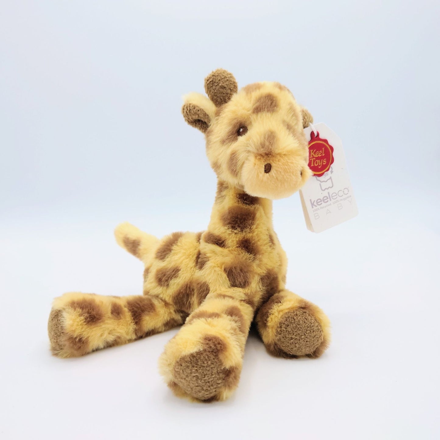 Plush toy baby giraffe from Keel toys sitting on a white surface with the legs splayed sideways towards the viewer. The white and red tag can be seen in the ear further away from the viewer.