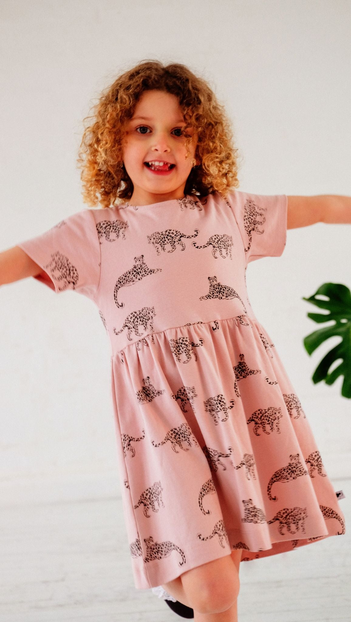 Lifestyle shot of girlwearing the leopard dress. She has curly hair and is holding her arms out to either side, facing the camera.
