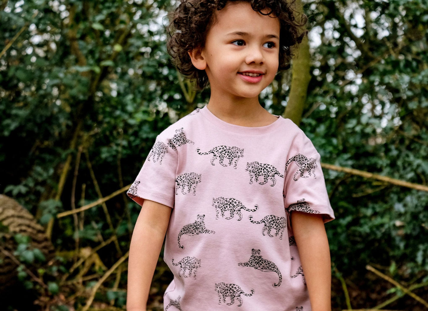 Lifestyle shot of child wearing the t-shirt standing in front of a dark green shrub.