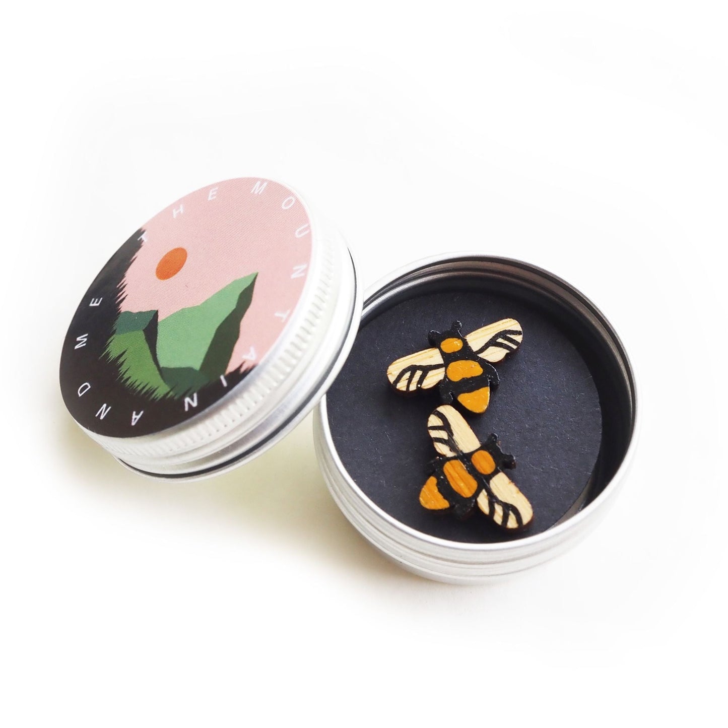 The Manchester bee earrings inside the screw top tin. The lid is resting against the side of the open tin and has the mountain and me branding on it.