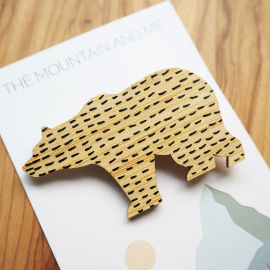 Close up of the bamboo bear brooch on the backing card that has The Mountain and Me branding and text. The bear has black short lines in a horizontal pattern all across it.